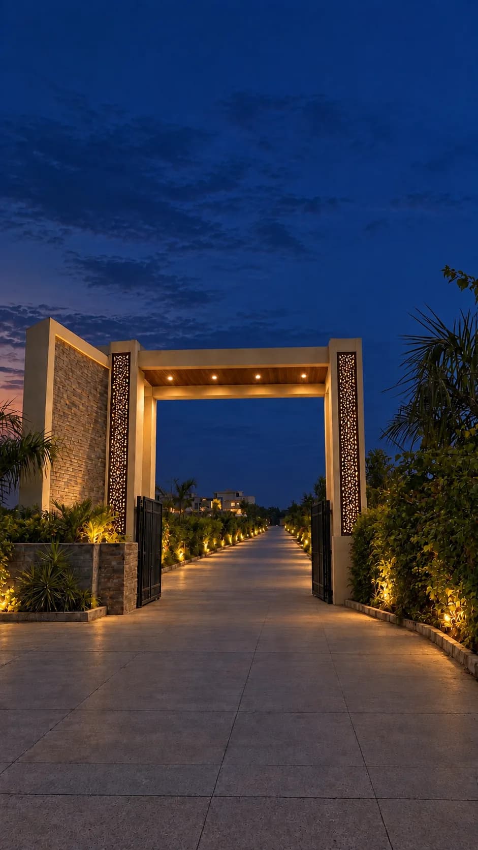 The entrance gate at dusk: two stone piers with lit jaali screens and a beam across, and the lamp-lined road into the estate